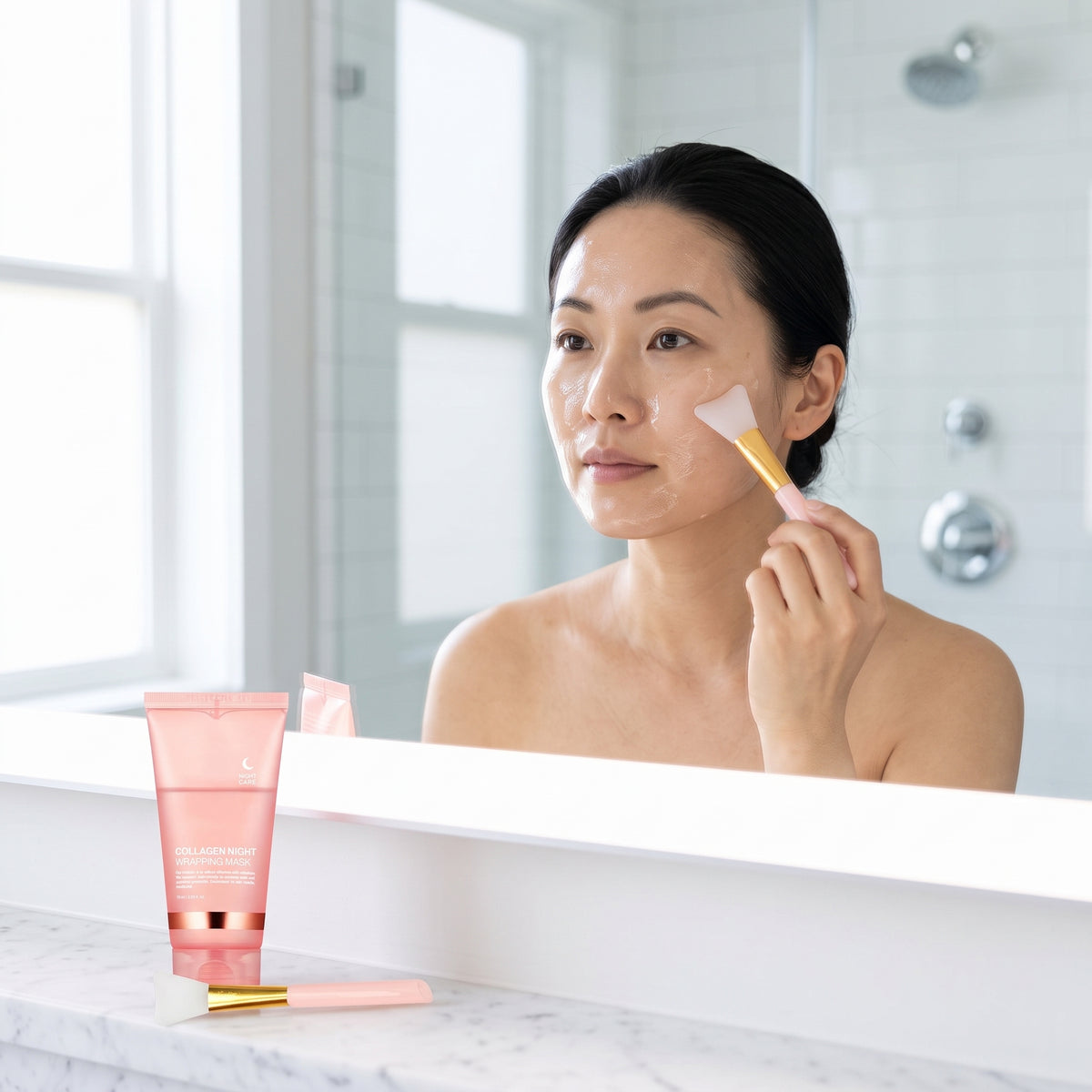lifestyle: A NEW photorealistic photograph composed from scratch. A female model, East Asian, in her 30s, with her hair pulled back, is shown from the chest up, applying the product mask to her cheek with an applicator brush. She is looking into a brightly lit mirror (out of frame). The background features a clean, white tiled modern bathroom with subtle chrome fixtures. Soft, even light emanates from a large window to the left, creating gentle highlights on her skin. The product tube and its applicator br…