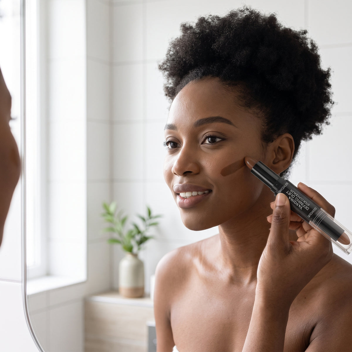 lifestyle: A NEW photorealistic photograph composed from scratch. A Black woman in her 30s with natural makeup is captured in a bright, modern bathroom. She is looking into a mirror (out of frame) and applying the product to her cheekbone. The background features a clean white tiled wall and a sleek, minimalist vanity with a simple plant in a ceramic pot. The product is held in her right hand, angled against her face, occupying approximately one-fifth of the frame height on the right side. Photographed at…